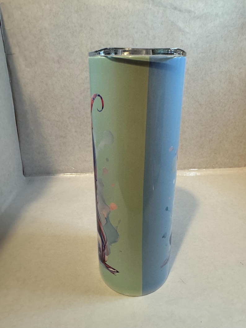 Może przedstawiać: Wysoki, izolowany kubek ze srebrną pokrywką. Kubek podzielony jest na dwie pionowe sekcje: jedną jasnozieloną i drugą jasno niebieską. Zielona sekcja zawiera kolorową ilustrację z r&oacute;żowymi i fioletowymi akcentami.