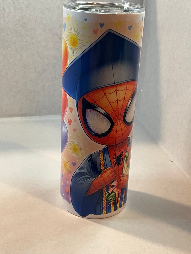 Pode incluir: Um copo de a&ccedil;o inoxid&aacute;vel branco com um design colorido que apresenta uma imagem de desenho animado do Homem-Aranha usando um chap&eacute;u de formatura e uma beca. O design tamb&eacute;m inclui bal&otilde;es coloridos, cora&ccedil;&otilde;es e estrelas.