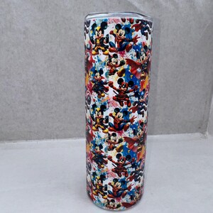 May include: A cylindrical stainless steel tumbler featuring a vibrant, multi-colored graphic print of various cartoon characters dressed as superheroes. The design includes splashes of blue, red, and yellow, with a metallic lid visible at the top.