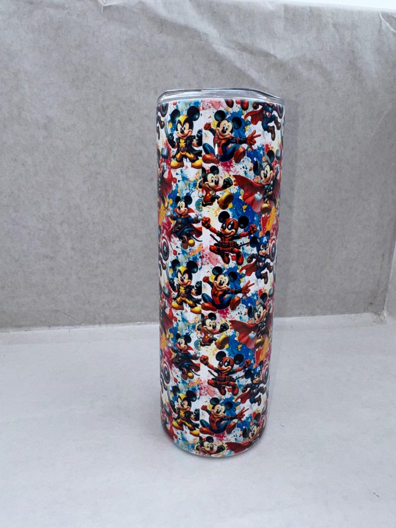 May include: A cylindrical stainless steel tumbler featuring a vibrant, multi-colored graphic print of various cartoon characters dressed as superheroes. The design includes splashes of blue, red, and yellow, with a metallic lid visible at the top.