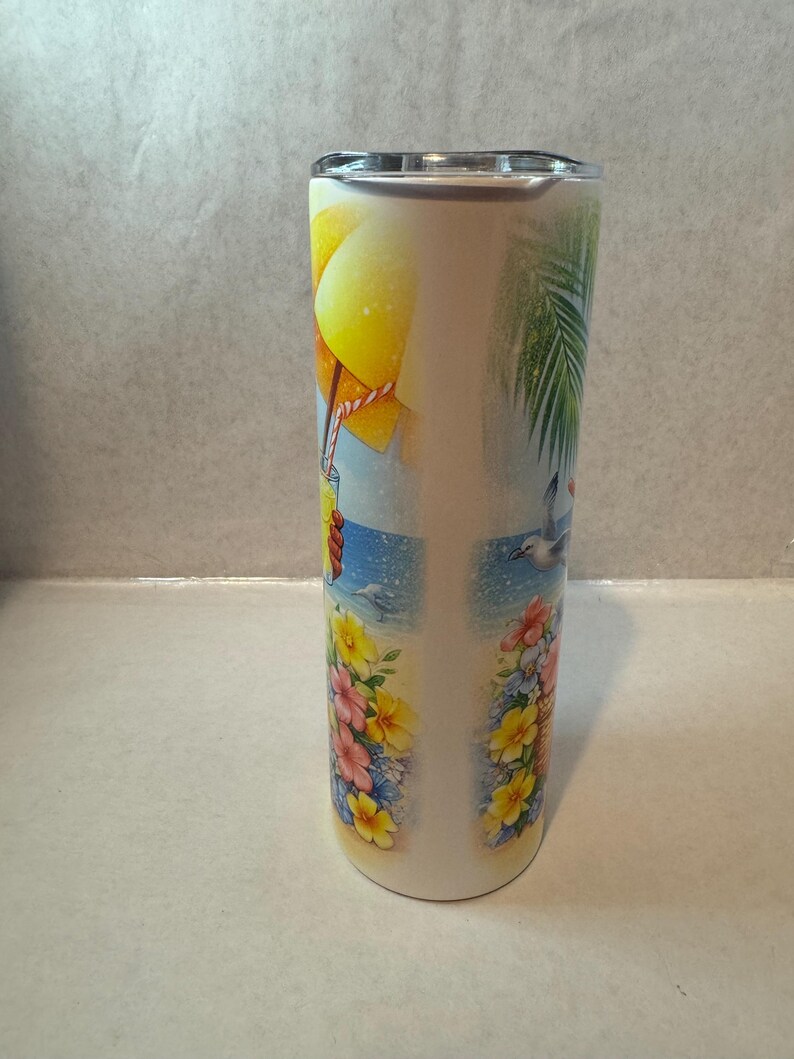 May include: A white stainless steel tumbler with a beach scene design. The design features a yellow umbrella, a blue ocean, a palm tree, and flowers.