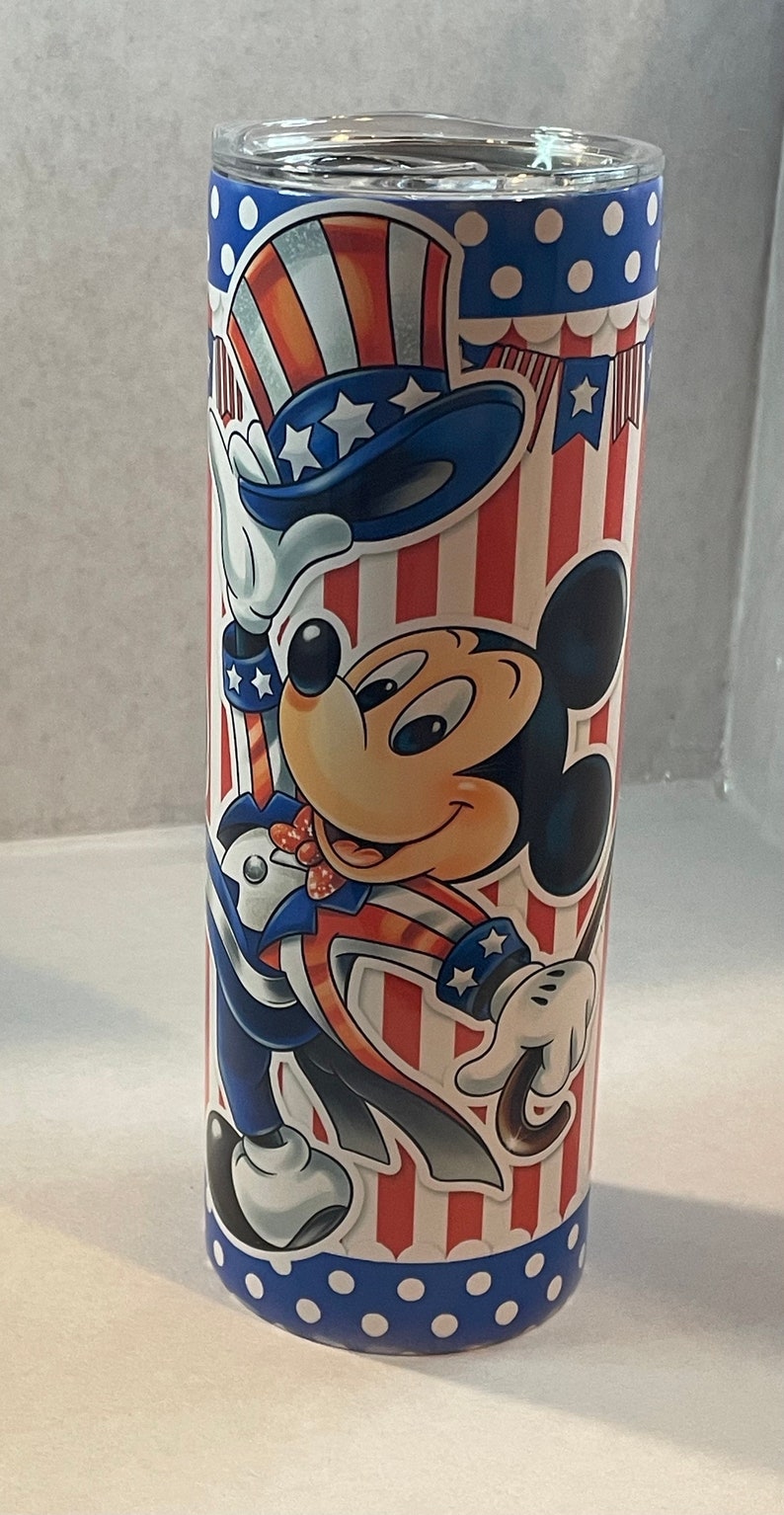 Peut inclure: Un gobelet en acier inoxydable avec un motif patriotique rouge, blanc et bleu repr&eacute;sentant Mickey Mouse habill&eacute; en Oncle Sam. Le gobelet a un couvercle blanc.