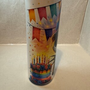 Może przedstawiać: Biały kubek ze stali nierdzewnej z kolorowym wzorem tortu urodzinowego i dekoracji imprezowych. Tort ma tęczowe warstwy, czarny lukier i zapalone świeczki. Wz&oacute;r zawiera r&oacute;wnież kolorowe flagi i gwiazdki.