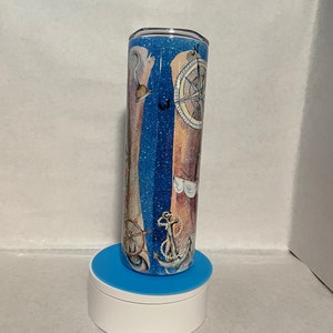 May include: A blue and white glitter tumbler with a nautical theme. The tumbler features a compass, anchor, and ship's wheel. The tumbler is on a blue and white stand.