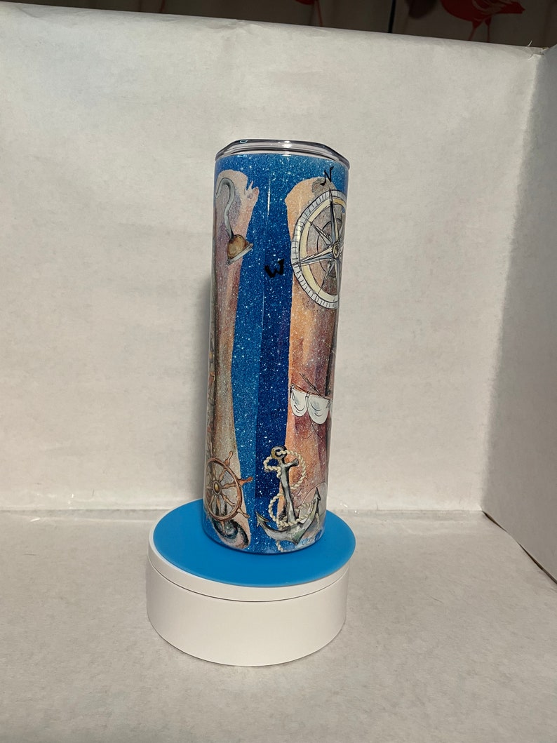 May include: A blue and white glitter tumbler with a nautical theme. The tumbler features a compass, anchor, and ship's wheel. The tumbler is on a blue and white stand.