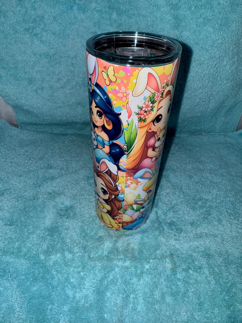 May include: A stainless steel tumbler with a black lid, decorated with cartoon characters in Easter bunny costumes. The wrap-around design features princesses and Easter-themed graphics. The background is a light blue.