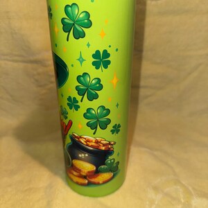 Może przedstawiać: Zielony kubek z czarną pokrywką, ozdobiony koniczyną, złotymi monetami i garnkiem złota. Projekt zawiera zielone koniczyny, złote monety i garnek złota, ze złotymi gwiazdami i diamentami. Kubek jest cylindryczny i wydaje się być wykonany z metalu.