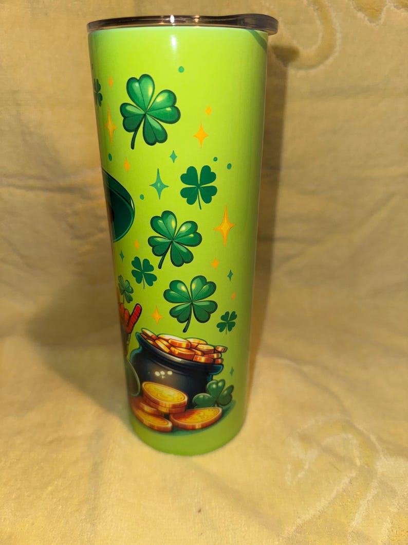 Może przedstawiać: Zielony kubek z czarną pokrywką, ozdobiony koniczyną, złotymi monetami i garnkiem złota. Projekt zawiera zielone koniczyny, złote monety i garnek złota, ze złotymi gwiazdami i diamentami. Kubek jest cylindryczny i wydaje się być wykonany z metalu.