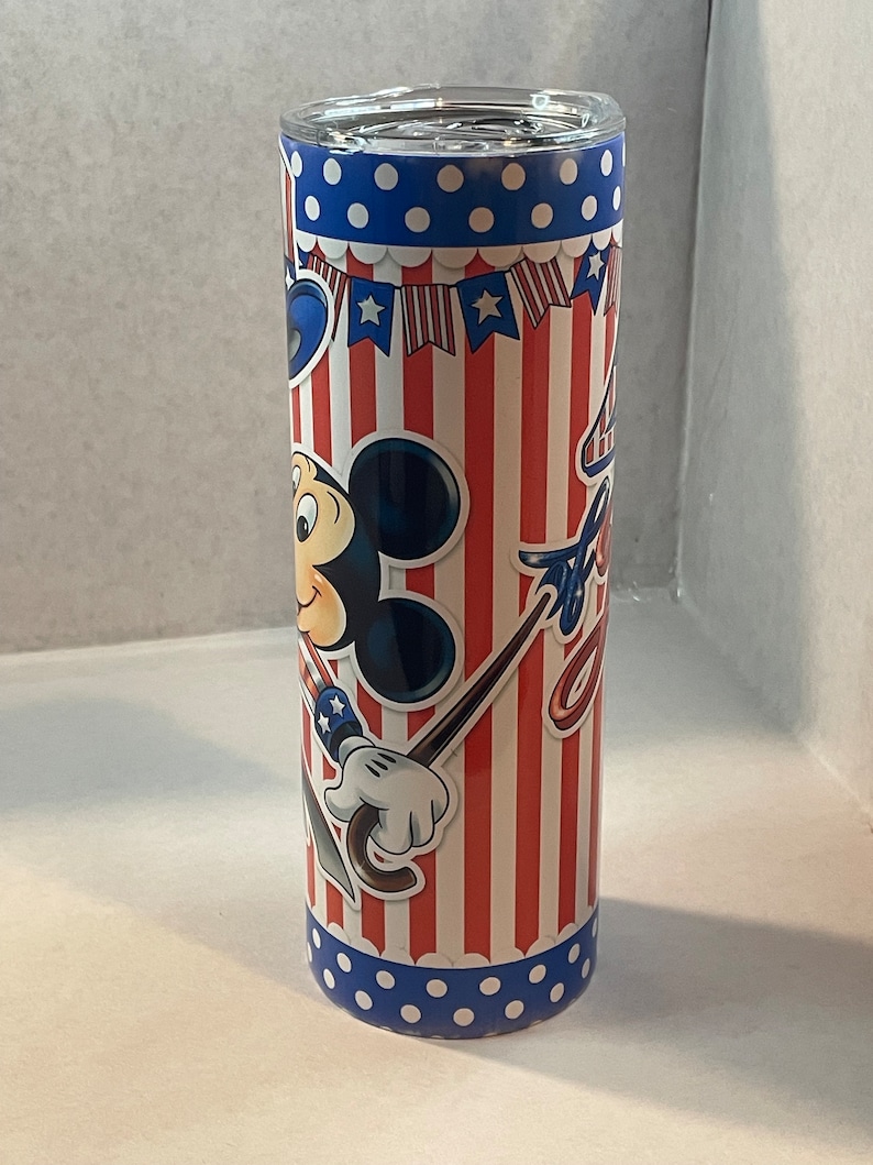 Peut inclure: Un gobelet isotherme rouge, blanc et bleu avec un motif Mickey Mouse. Le gobelet pr&eacute;sente un fond ray&eacute; rouge et blanc avec des pois bleus en haut et en bas. Mickey Mouse tient un parapluie rouge, blanc et bleu et porte un chapeau rouge, blanc et bleu.