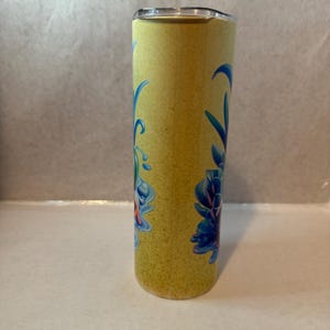 K&ouml;nnte beinhalten: Ein gelber Becher mit einem floralen Design in Blau und T&uuml;rkis. Der Edelstahlbecher hat einen Deckel und eine strukturierte Oberfl&auml;che. Das florale Design befindet sich auf der Vorderseite des Bechers.