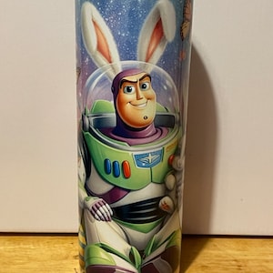 Puede incluir: Un vaso cil&iacute;ndrico con Buzz Lightyear con un traje espacial y orejas de conejo. El dise&ntilde;o incluye un fondo estrellado, mariposas y Buzz sonriendo. El vaso es una vibrante mezcla de verde, morado y blanco.