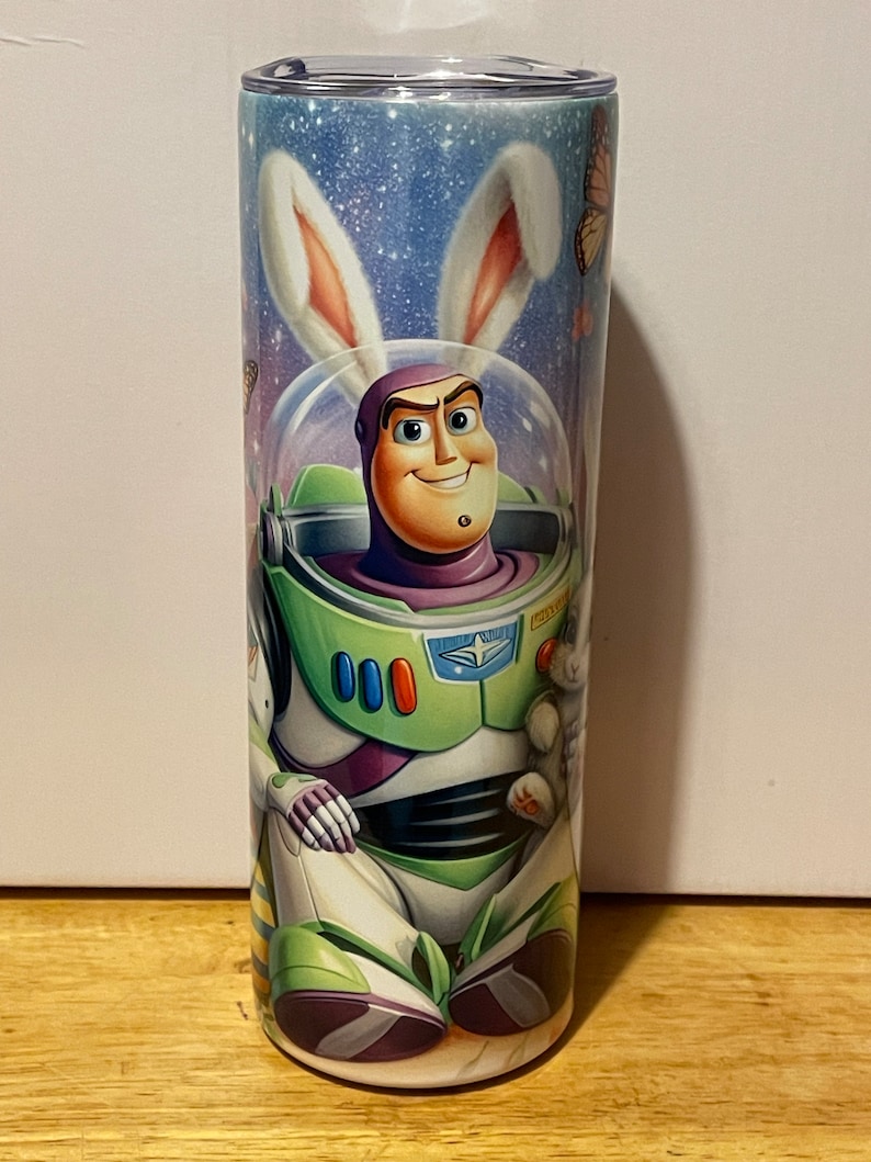 Puede incluir: Un vaso cil&iacute;ndrico con Buzz Lightyear con un traje espacial y orejas de conejo. El dise&ntilde;o incluye un fondo estrellado, mariposas y Buzz sonriendo. El vaso es una vibrante mezcla de verde, morado y blanco.