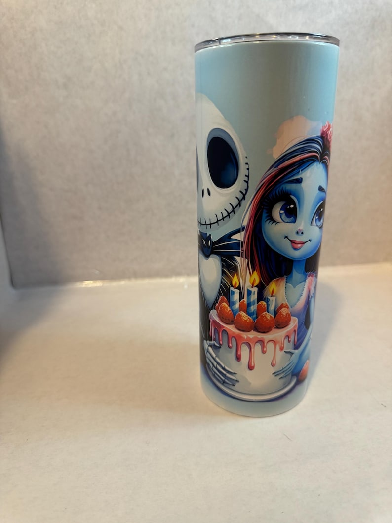 Może przedstawiać: Wysoki, cylindryczny kubek z kresk&oacute;wkową ilustracją Jacka Skellingtona i Sally z The Nightmare Before Christmas. Sally trzyma tort ze świecami i truskawkami. Tło jest jasnoniebieskie.