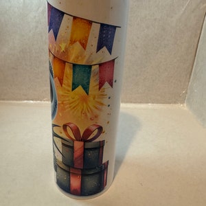 Może przedstawiać: Biały kubek ze stali nierdzewnej z kolorowym wzorem na przyjęcie urodzinowe. Wz&oacute;r przedstawia niebieską postać z czerwoną kokardką, kolorowe flagi i fajerwerki. Kubek ma czarną pokrywkę.