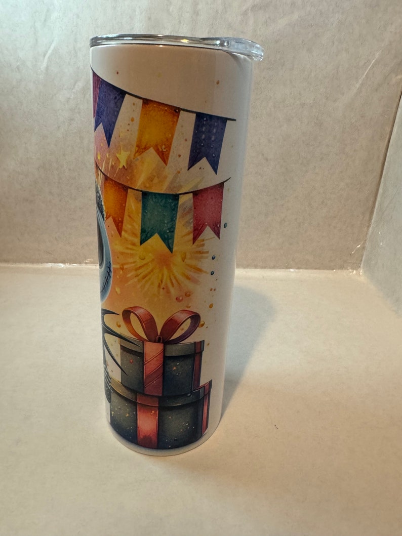 Może przedstawiać: Biały kubek ze stali nierdzewnej z kolorowym wzorem na przyjęcie urodzinowe. Wz&oacute;r przedstawia niebieską postać z czerwoną kokardką, kolorowe flagi i fajerwerki. Kubek ma czarną pokrywkę.