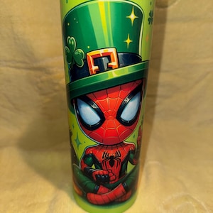 Może przedstawiać: Zielony kubek z postacią z kresk&oacute;wki Spider-Man w zielonym kapeluszu krasnoludka ze złotą klamrą i koniczyną. Kubek ma błyszczące metaliczne wykończenie i jest ozdobiony gwiazdami.