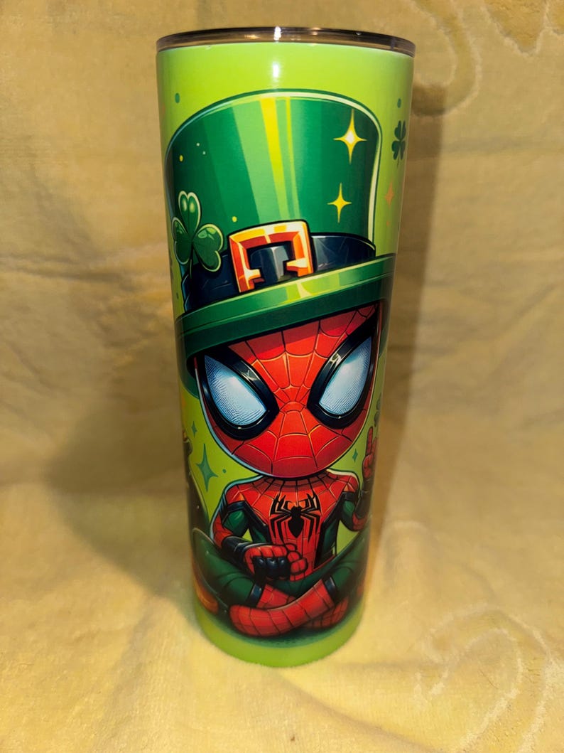 Może przedstawiać: Zielony kubek z postacią z kresk&oacute;wki Spider-Man w zielonym kapeluszu krasnoludka ze złotą klamrą i koniczyną. Kubek ma błyszczące metaliczne wykończenie i jest ozdobiony gwiazdami.
