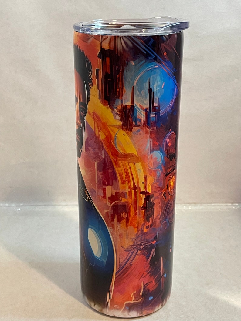May include: A stainless steel tumbler with a colorful abstract design featuring a superhero character. The design includes a cityscape, a blue sky, and a yellow and orange sunset.