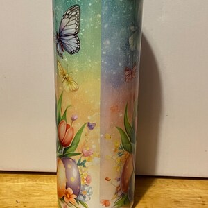 Puede incluir: Un vaso alto y aislado con un dise&ntilde;o de mariposas y flores. El vaso presenta un fondo degradado de azul, verde y amarillo. Mariposas y flores est&aacute;n impresas en el vaso, con una tapa en la parte superior.