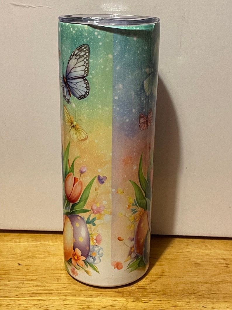 Puede incluir: Un vaso alto y aislado con un dise&ntilde;o de mariposas y flores. El vaso presenta un fondo degradado de azul, verde y amarillo. Mariposas y flores est&aacute;n impresas en el vaso, con una tapa en la parte superior.