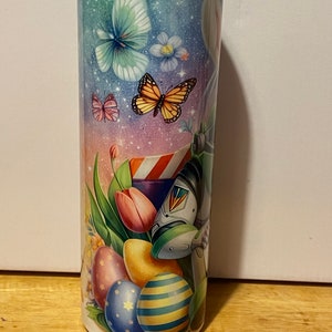 Puede incluir: Un vaso de acero inoxidable con un dise&ntilde;o colorido con tem&aacute;tica de Pascua. El dise&ntilde;o presenta mariposas, flores, huevos de Pascua y tulipanes sobre un fondo pastel. El vaso tiene una tapa de pl&aacute;stico transparente.