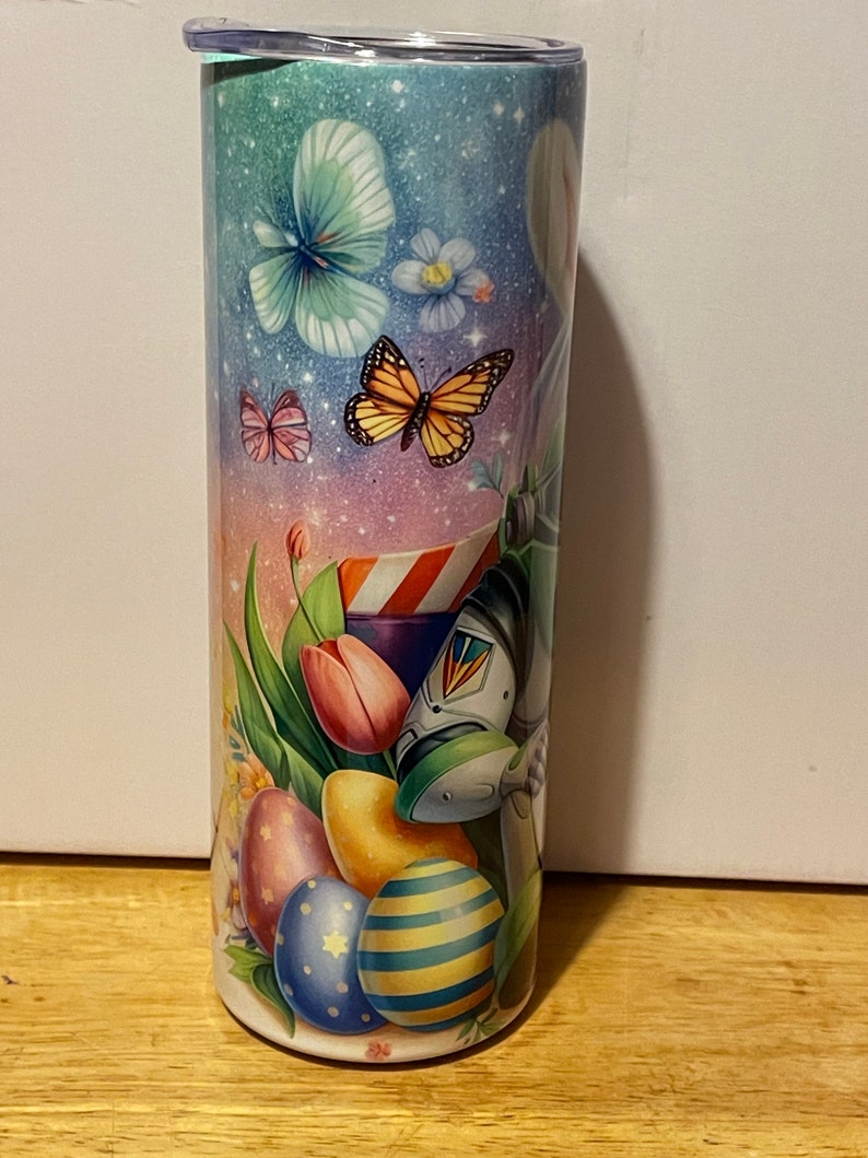 Puede incluir: Un vaso de acero inoxidable con un dise&ntilde;o colorido con tem&aacute;tica de Pascua. El dise&ntilde;o presenta mariposas, flores, huevos de Pascua y tulipanes sobre un fondo pastel. El vaso tiene una tapa de pl&aacute;stico transparente.