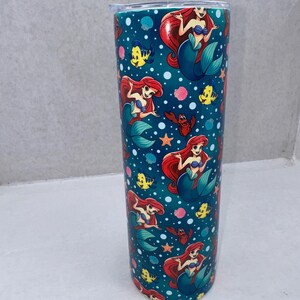 May include: A cylindrical tumbler featuring a repeating pattern of a red-haired mermaid, a yellow fish, and a red crab against a dark teal background filled with white bubbles, pink coral, and orange starfish, topped with a clear plastic lid.