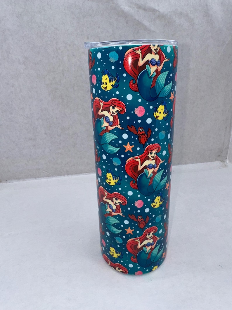 May include: A cylindrical tumbler featuring a repeating pattern of a red-haired mermaid, a yellow fish, and a red crab against a dark teal background filled with white bubbles, pink coral, and orange starfish, topped with a clear plastic lid.