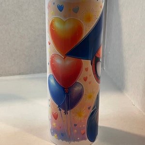 Pode incluir: Um copo de a&ccedil;o inoxid&aacute;vel branco com um design colorido que apresenta um super-her&oacute;i de desenho animado usando um chap&eacute;u de formatura, bal&otilde;es vermelhos, amarelos e azuis e cora&ccedil;&otilde;es.