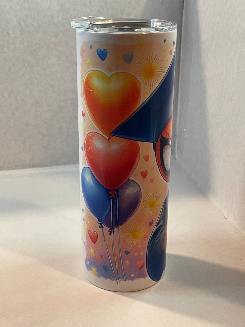 Pode incluir: Um copo de a&ccedil;o inoxid&aacute;vel branco com um design colorido que apresenta um super-her&oacute;i de desenho animado usando um chap&eacute;u de formatura, bal&otilde;es vermelhos, amarelos e azuis e cora&ccedil;&otilde;es.