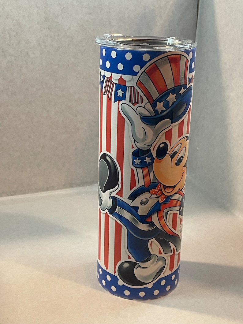Peut inclure: Un gobelet en acier inoxydable avec un motif patriotique repr&eacute;sentant Mickey Mouse habill&eacute; en Oncle Sam. Le gobelet a un fond ray&eacute; rouge, blanc et bleu avec des pois blancs en haut et en bas.
