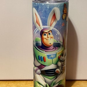 Puede incluir: Un vaso cil&iacute;ndrico alto con una ilustraci&oacute;n de dibujos animados de Buzz Lightyear con un traje espacial y orejas de conejo. El dise&ntilde;o incluye un fondo de cielo estrellado, una mariposa y un conejito.