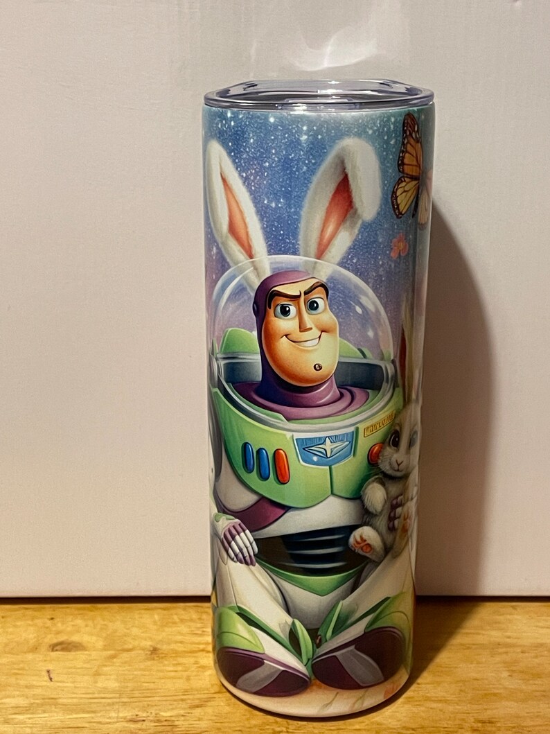 Puede incluir: Un vaso cil&iacute;ndrico alto con una ilustraci&oacute;n de dibujos animados de Buzz Lightyear con un traje espacial y orejas de conejo. El dise&ntilde;o incluye un fondo de cielo estrellado, una mariposa y un conejito.