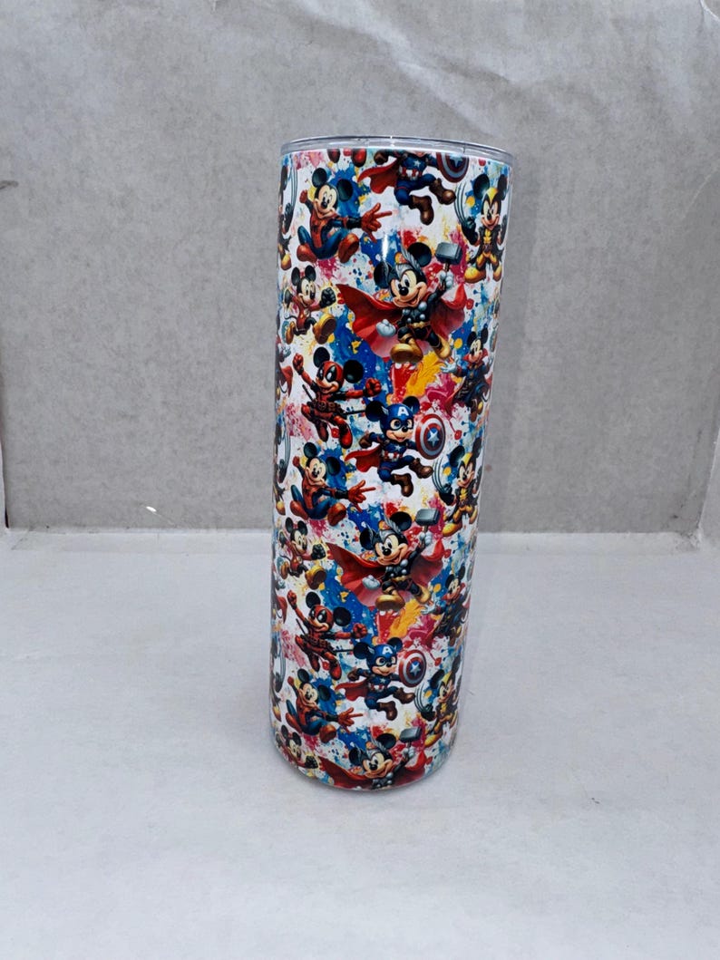 May include: A cylindrical stainless steel tumbler featuring a repeating pattern of cartoon mice dressed as various superheroes. The design includes vibrant splashes of blue, red, and yellow paint against a white background, with a metallic rim at the top.