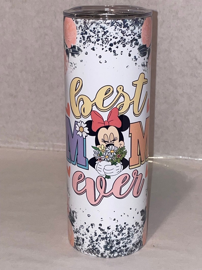 Puede incluir: Un vaso blanco con un fondo moteado en blanco y negro. El vaso tiene un gr&aacute;fico rosa, amarillo y morado que dice "best mom ever" con una imagen de dibujos animados de Minnie Mouse sosteniendo un ramo de flores.