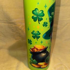 Może przedstawiać: Zielony kubek ze stali nierdzewnej z motywem Dnia Świętego Patryka. Projekt zawiera kapelusz krasnoluda, koniczyny, złote monety i garnek złota. Kubek ma srebrną pokrywę i ma około 20 cm wysokości.
