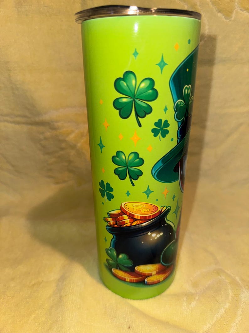 Może przedstawiać: Zielony kubek ze stali nierdzewnej z motywem Dnia Świętego Patryka. Projekt zawiera kapelusz krasnoluda, koniczyny, złote monety i garnek złota. Kubek ma srebrną pokrywę i ma około 20 cm wysokości.