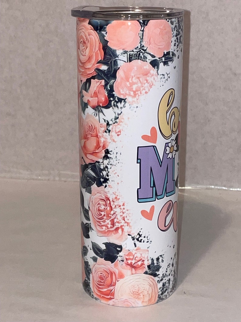 Puede incluir: Un vaso de acero inoxidable blanco con un dise&ntilde;o floral y el texto "Love Mom" en rosa, morado y amarillo. El vaso tiene una tapa negra.