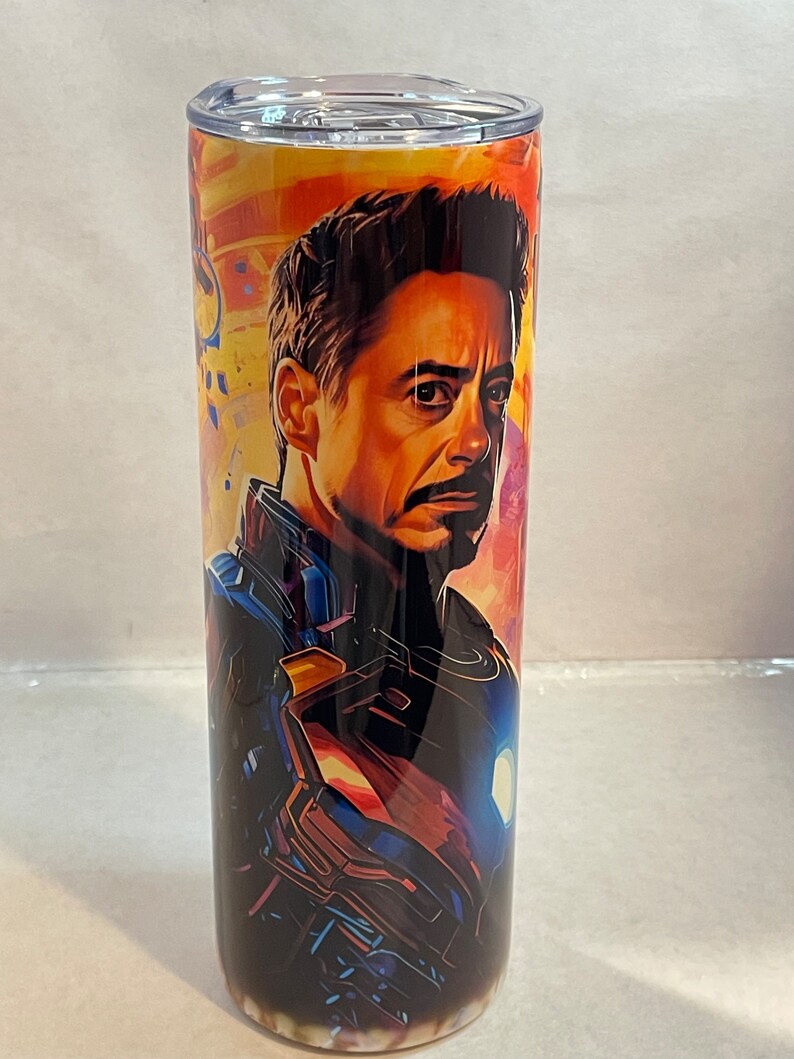 May include: A stainless steel tumbler with a colorful image of Iron Man in his suit. The background is a mix of orange, yellow, and blue.
