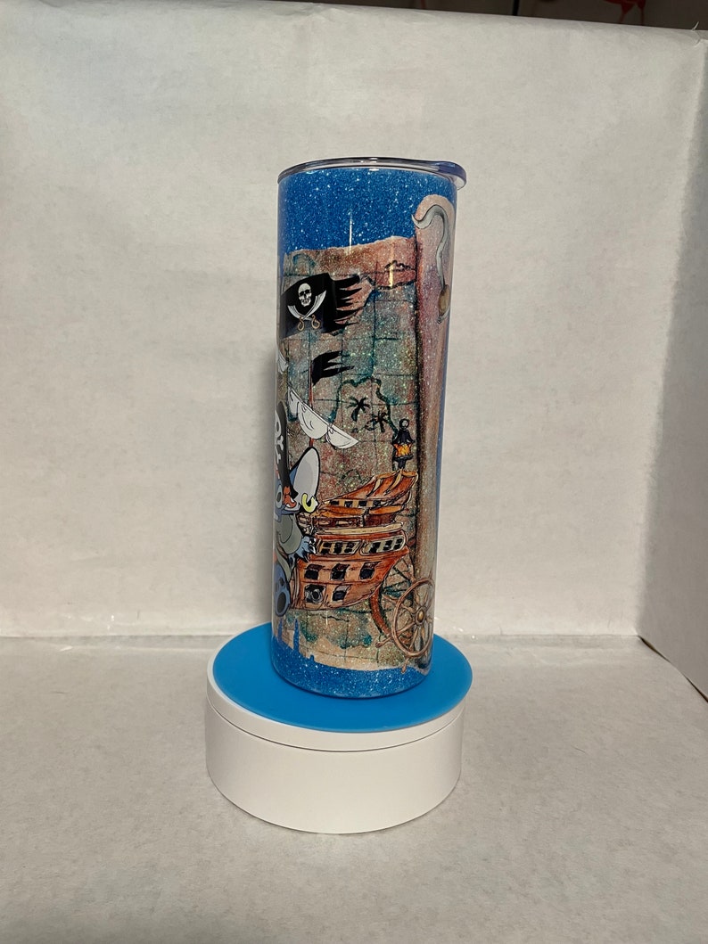 May include: A blue and white glitter tumbler with a pirate map design. The map features a pirate ship, a skull and crossbones, and a treasure chest.