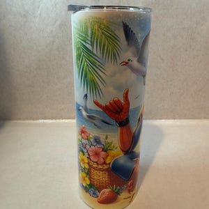 May include: A stainless steel tumbler with a colorful design featuring a cartoon superhero character on a beach with palm trees, flowers, and seagulls. The tumbler has a lid.