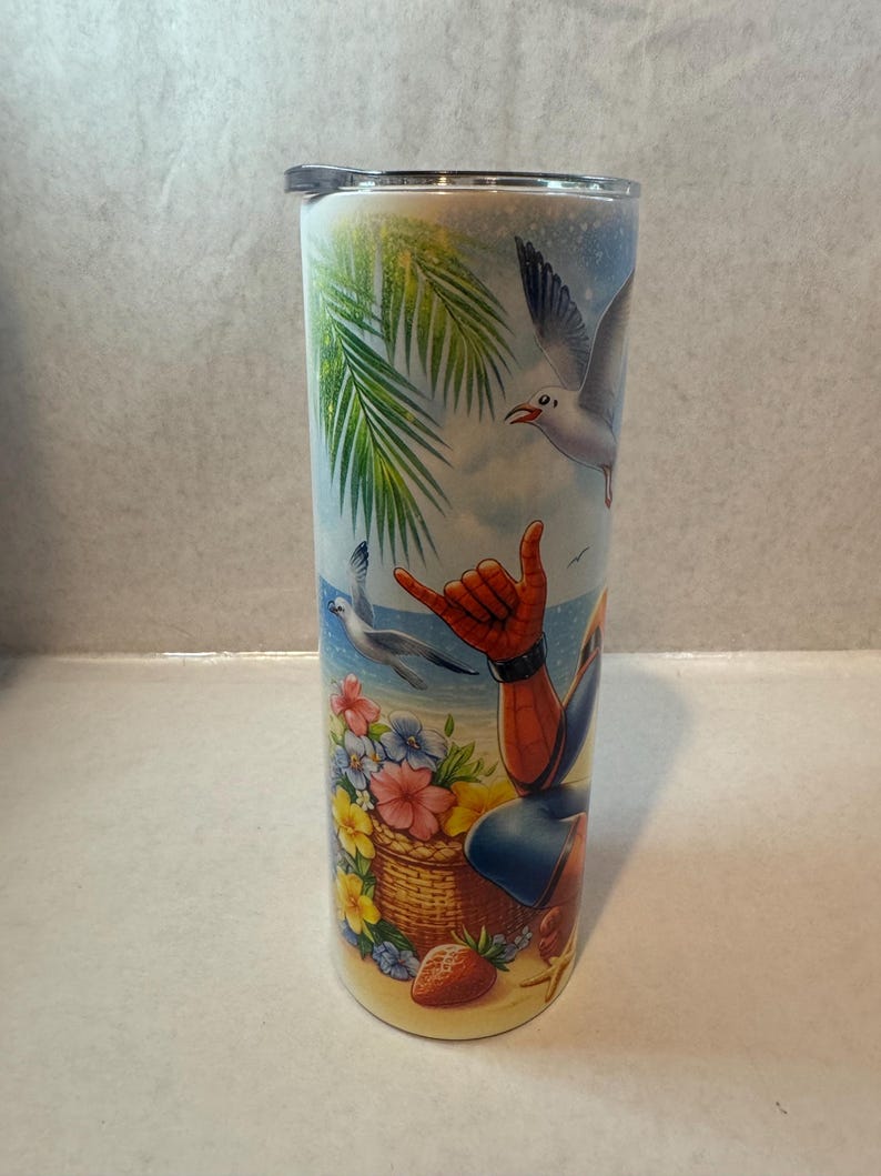 May include: A stainless steel tumbler with a colorful design featuring a cartoon superhero character on a beach with palm trees, flowers, and seagulls. The tumbler has a lid.