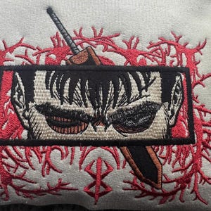 May include: Embroidered patch featuring a close-up of a character's face with a sword behind it. The character has black hair and a serious expression. The background is a red, branching design.