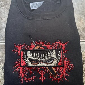 May include: Black sweatshirt with a red embroidered design featuring a character's eye and a sword. The design is surrounded by red thorns and a red symbol.