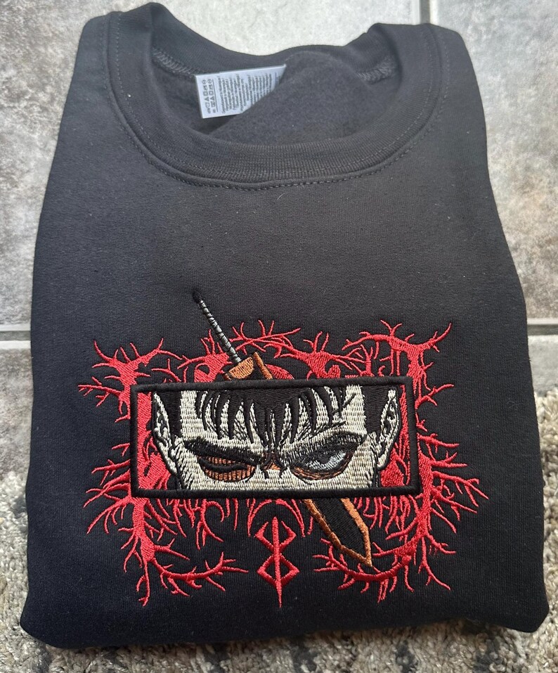May include: Black sweatshirt with a red embroidered design featuring a character's eye and a sword. The design is surrounded by red thorns and a red symbol.
