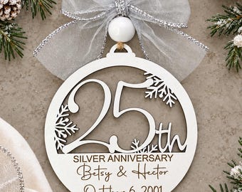 May include: A circular white wooden ornament featuring a cutout of the number 25 and snowflakes. The text reads SILVER ANNIVERSARY, Betsy & Hector, and October 6, 2001. It is topped with a white bead and a sheer silver ribbon with a jute twine loop.