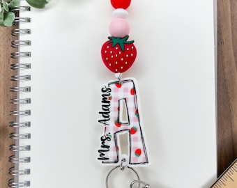 May include: A lanyard with a red strawberry charm and a clear acrylic letter "A" with a red gingham pattern and strawberry accents. The lanyard has pink, white, and red beads and the text "Mrs. Adams". Includes a silver-colored clasp and key ring.