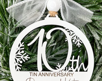 May include: A white circular ornament featuring a cutout number 10 and snowflake designs. The text reads TIN ANNIVERSARY, Ryan and Kate, and June 18, 2016. It is topped with a white bead and a sheer white ribbon bow attached to a jute twine loop.