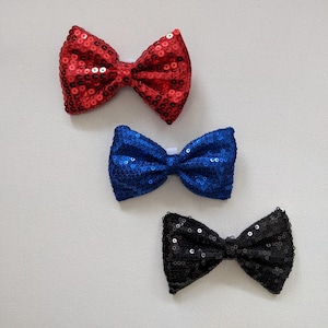 Può includere: Tre papillon con paillettes rossi, blu e neri. I papillon sono disposti su una superficie bianca. Ogni papillon ha una piccola fascia bianca al centro.