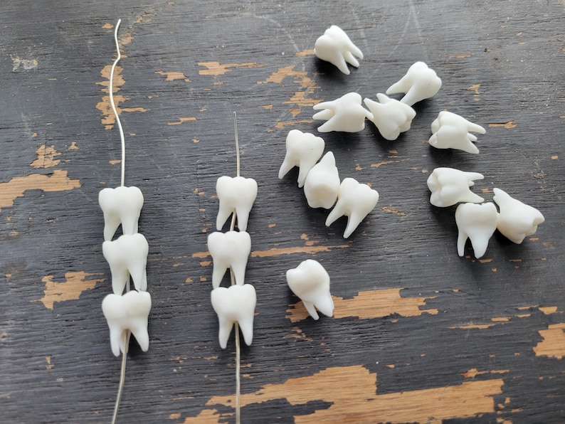 Pu&ograve; includere: Primo piano di denti di plastica bianca infilati su fili sottili. I denti sono disposti in uno schema di tre denti per filo, con due fili visibili. Ci sono anche diversi denti sciolti sparsi intorno ai fili.